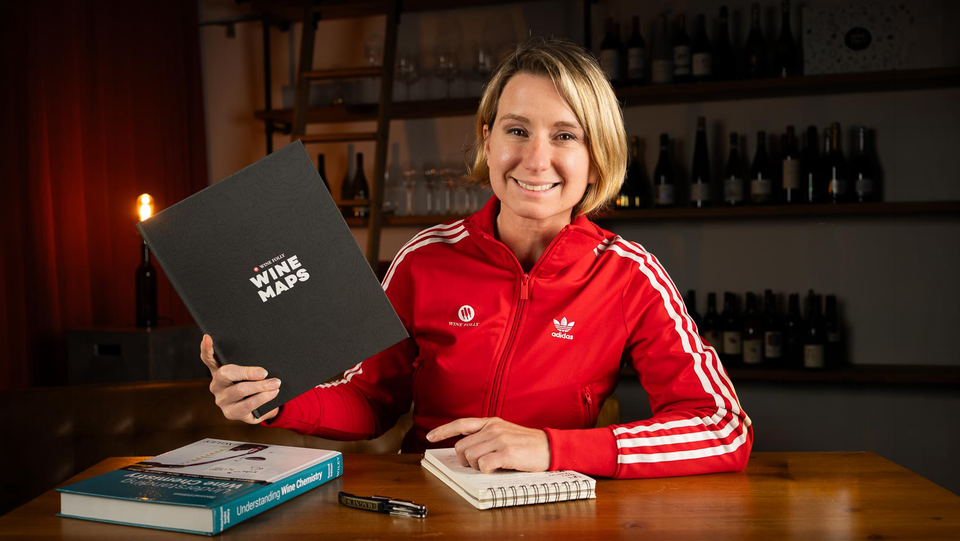 Madeline Puckette smiles at the camera in a red track jacket, holding up a dark hardcover book titled Wine Maps, seated at a wooden table with a notebook and books in a warmly lit room.