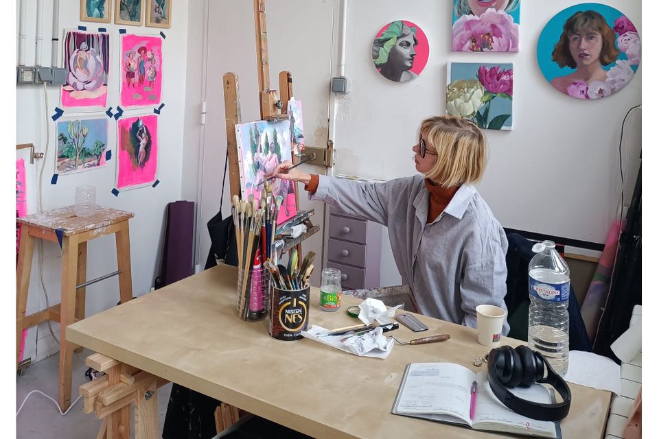 Liz Adams, a blonde woman in glasses and a striped shirt, paints at a wooden easel in a white studio with colourful canvases on the walls.