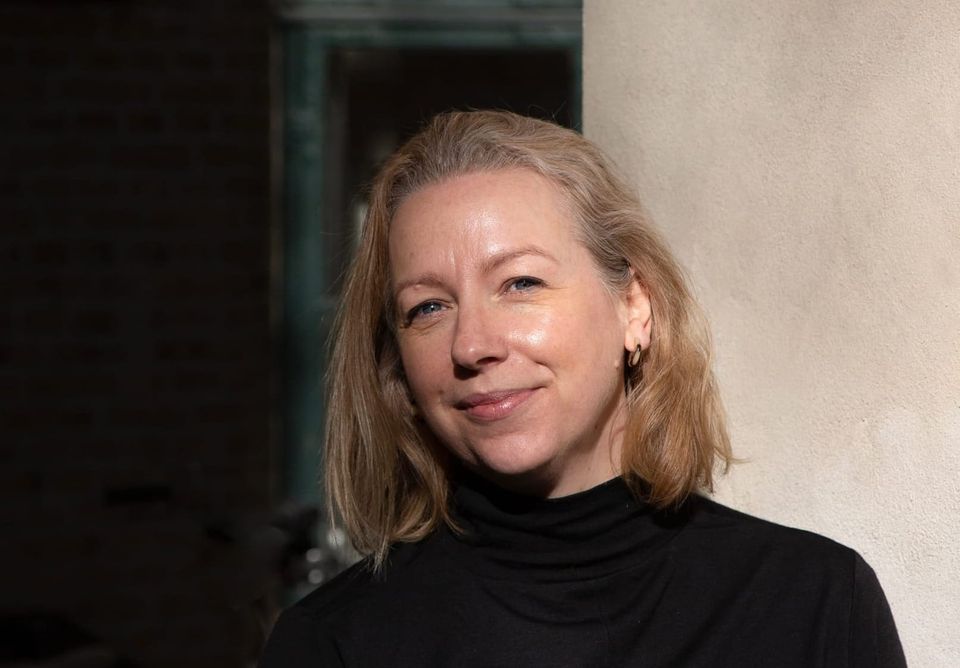 Laura Hall, a woman with shoulder-length blonde hair and a black turtleneck, smiles softly in warm sunlight against a cream wall.