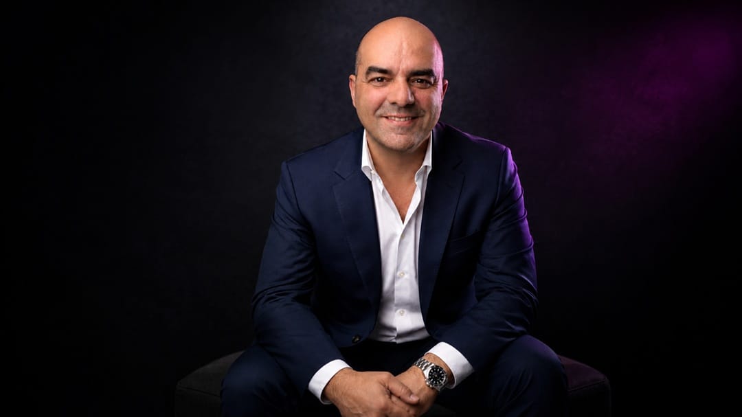 Fotografia do palestrante de vendas Leandro Branquinho. Ele está sentado, usando um traje formal e sorrindo para a câmera