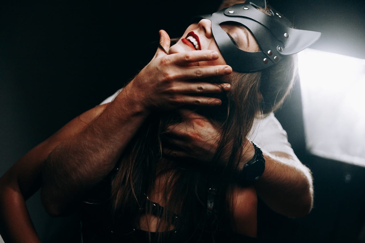 Masked woman held by the throat in a moment of consensual dominance, embodying the intensity and erotic thrill of power play explored in sensual choking scenes.