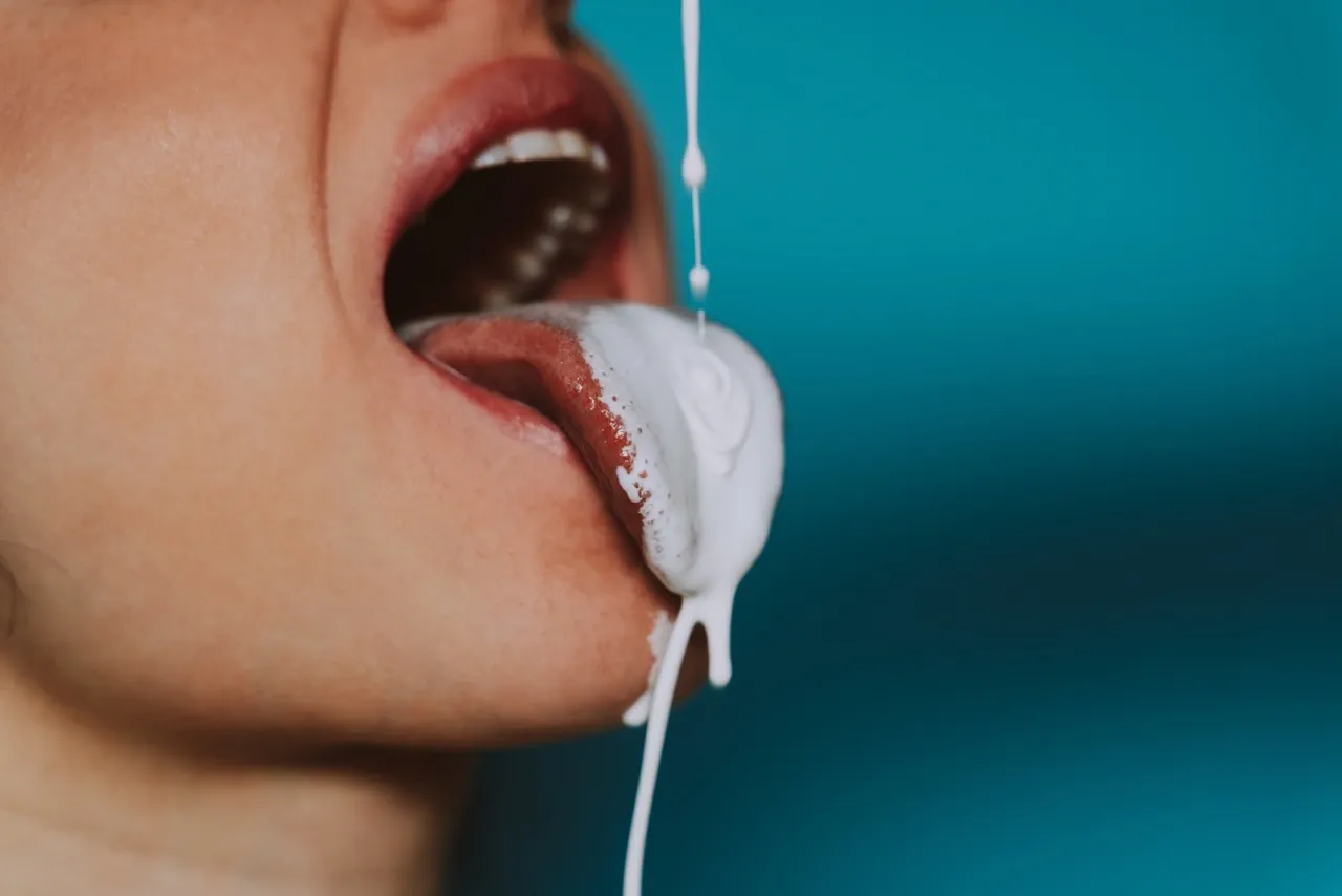 Mouth open wide as thick cream drips onto the tongue, representing playful sensuality and temptation.