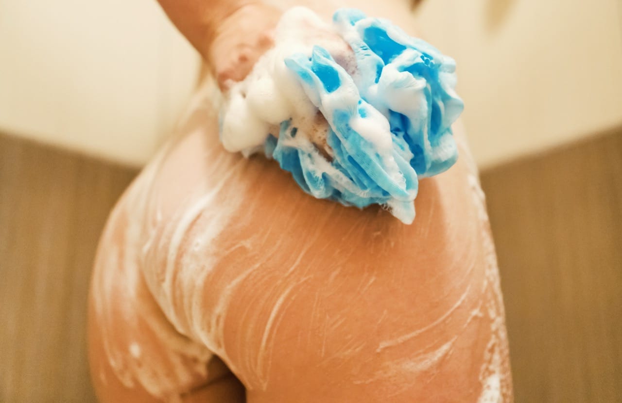 Blue bath sponge covered in soap gliding across a woman’s wet skin in the shower wiping off cum from her ass