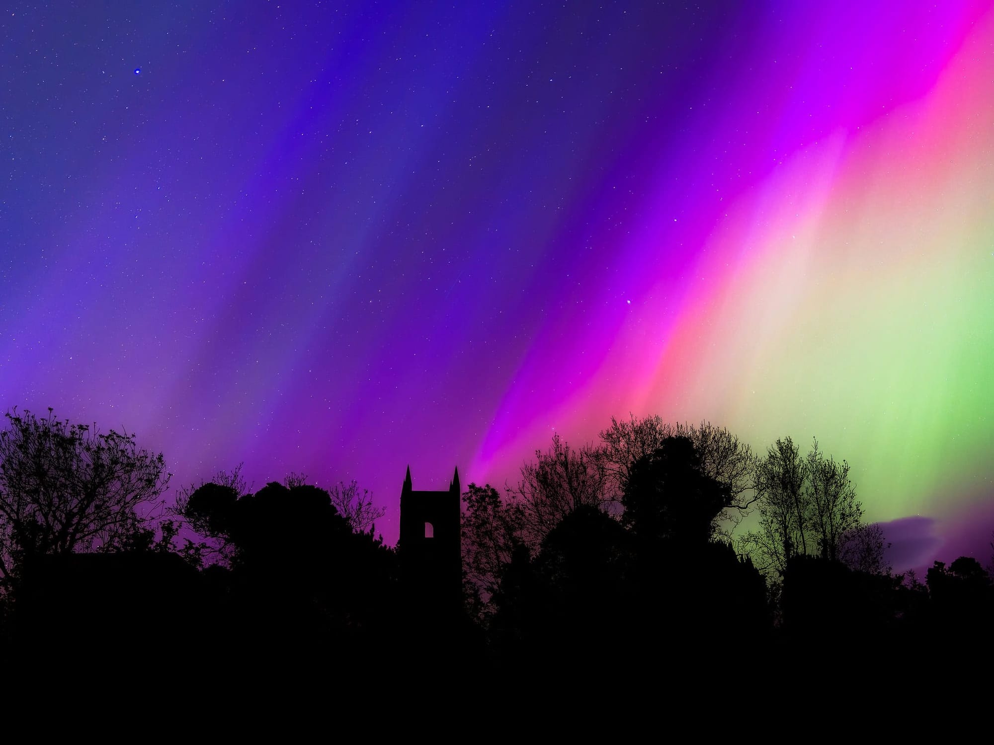 Dark ink blue, red, and green beams of light on a pitch black silhouette of trees and church tower.