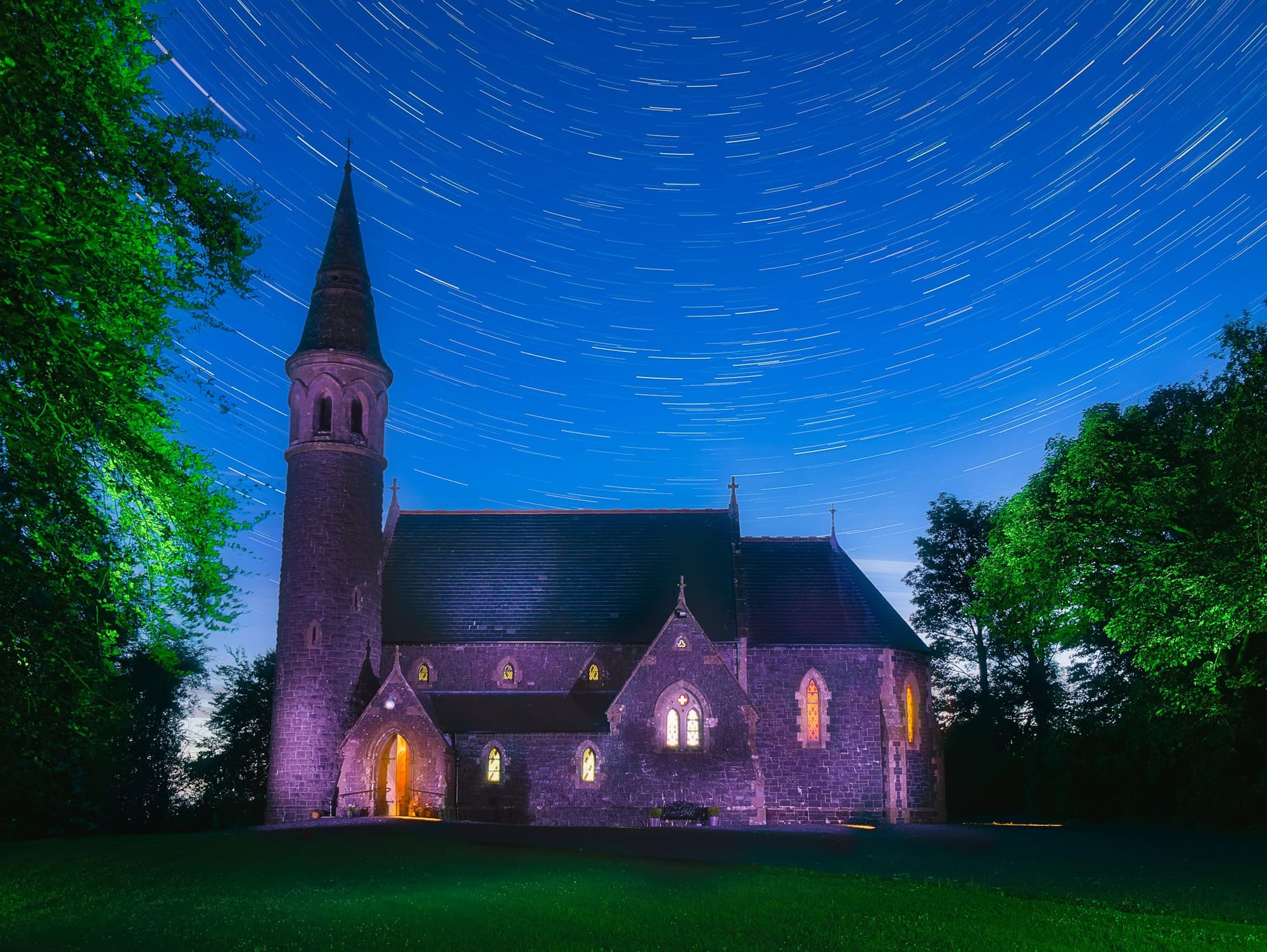 A stone church bathed in purple light, the windows and doorway glow in warmth, star trails above