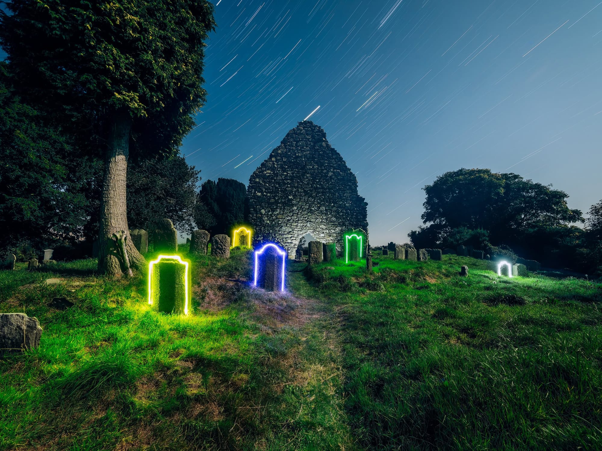 A moonlit church ruin, star trails in the sky, and neon light glows around the headstones
