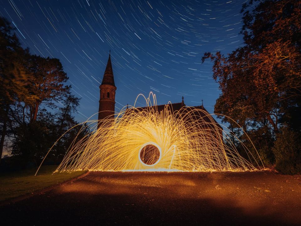 A long exposure featuring steel wool fire trails and star trails with an old church in the background 