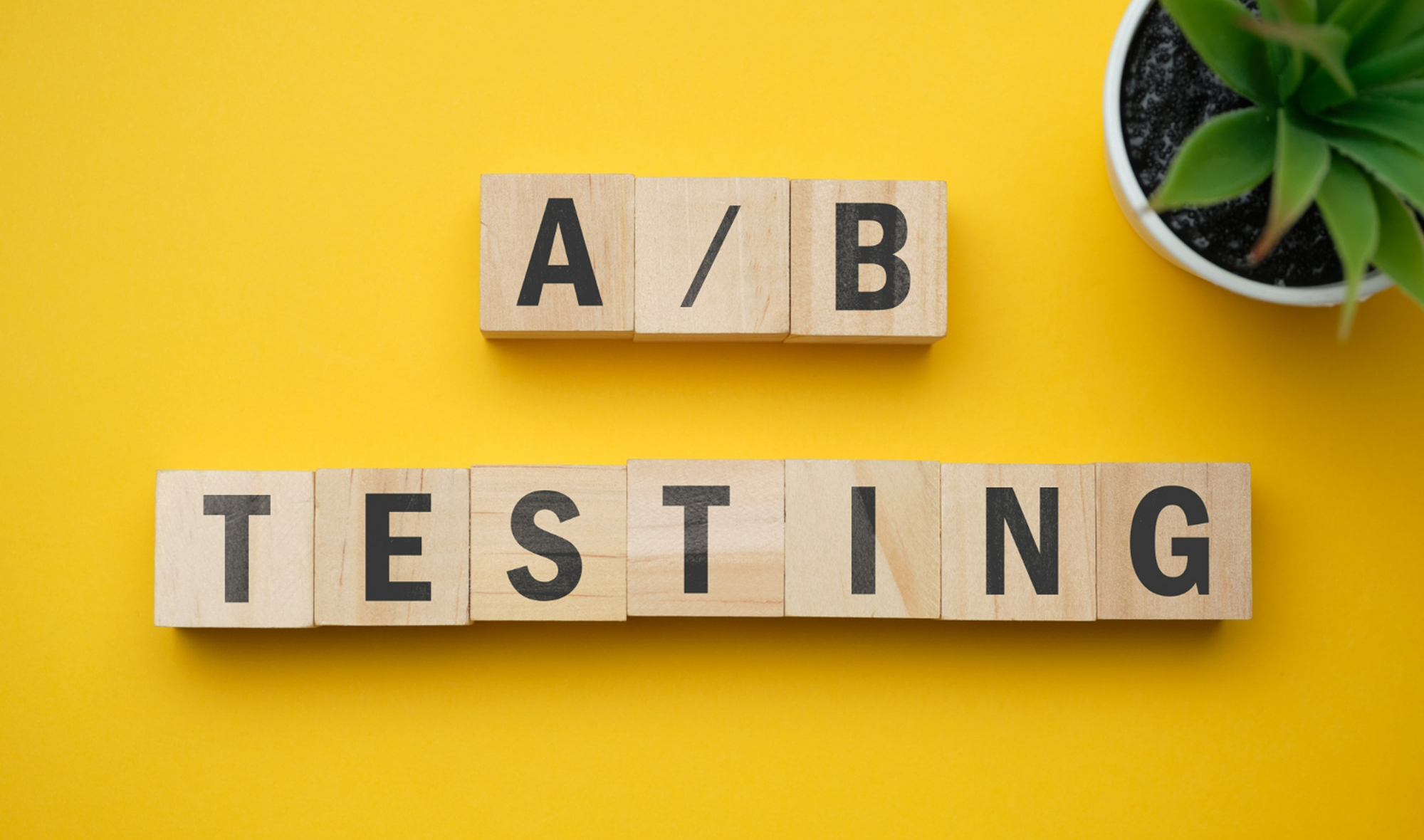A/B Testing이라는 단어가 우드 블록 위에 적혀 있다. 주위 배경은 노란색이고 오른쪽 상단에 화분이 하나 있다.