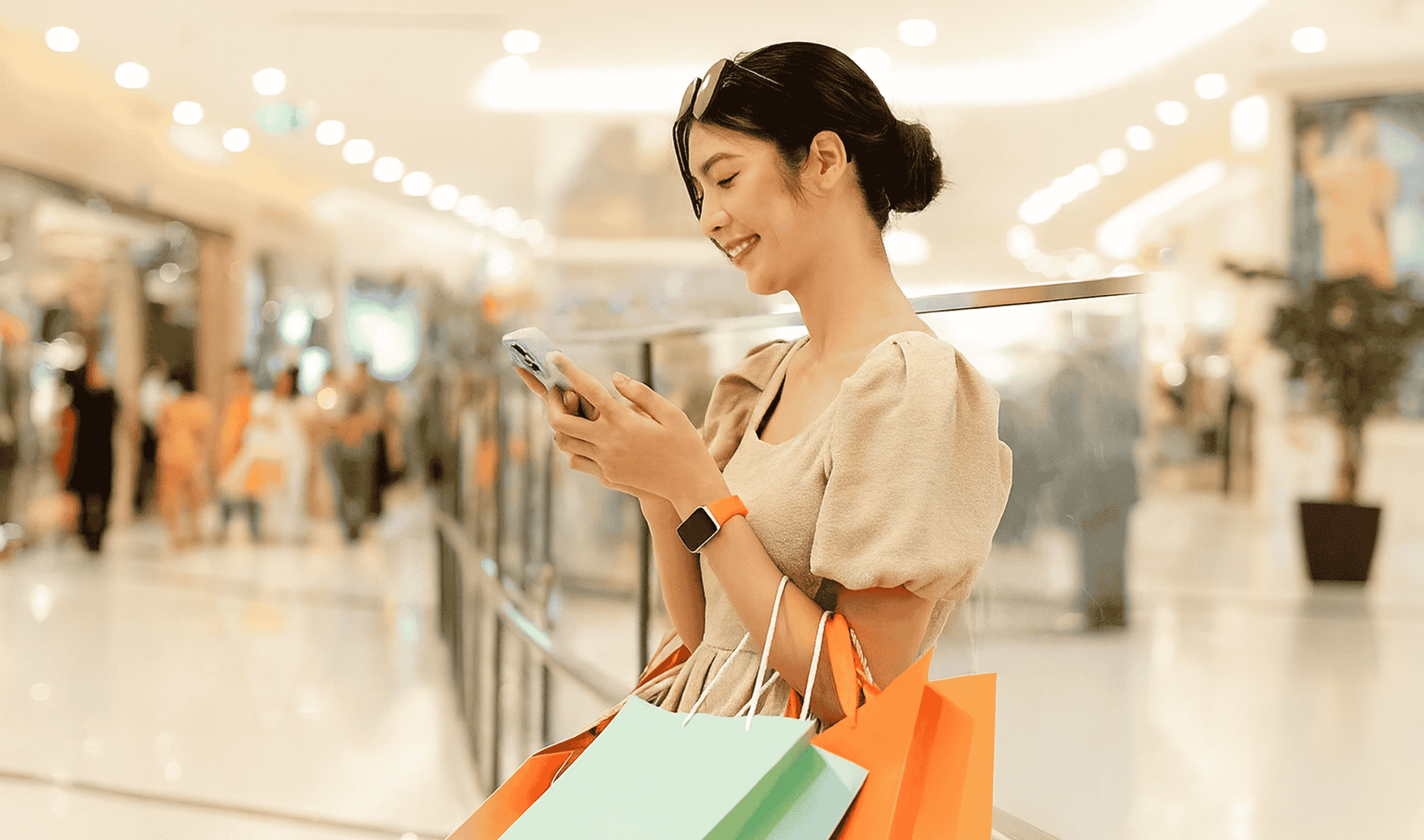 실내 쇼핑몰에서 쇼핑백을 팔에 걸친 채 스마트폰을 보면서 웃고 있는 여자