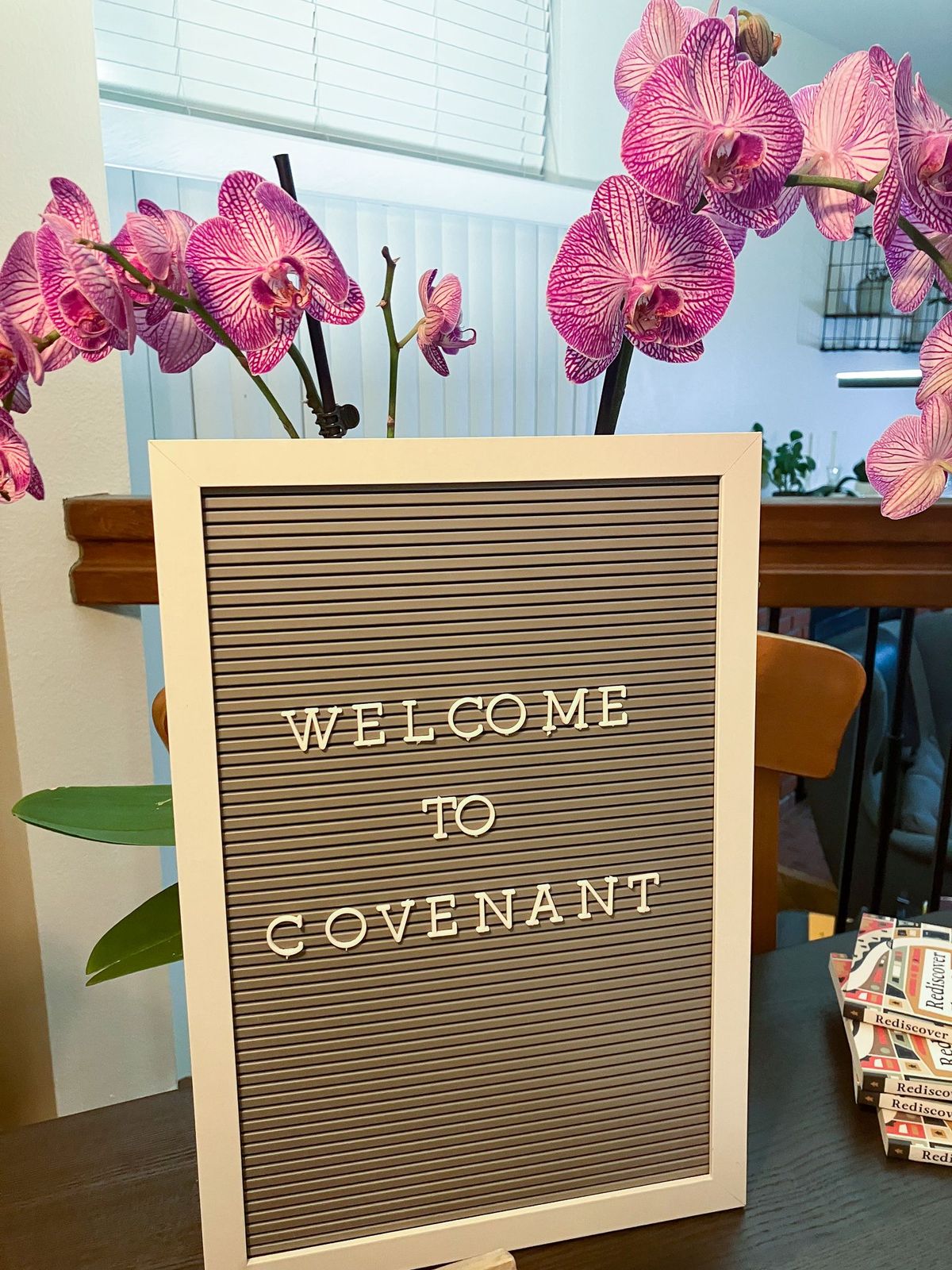 Sign on a table that says "WELCOME TO COVENANT" in front of orchid flowers.