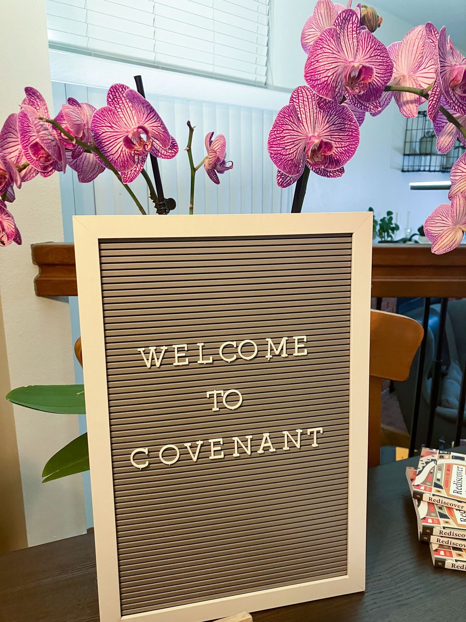 Sign on a table that says "WELCOME TO COVENANT" in front of orchid flowers.