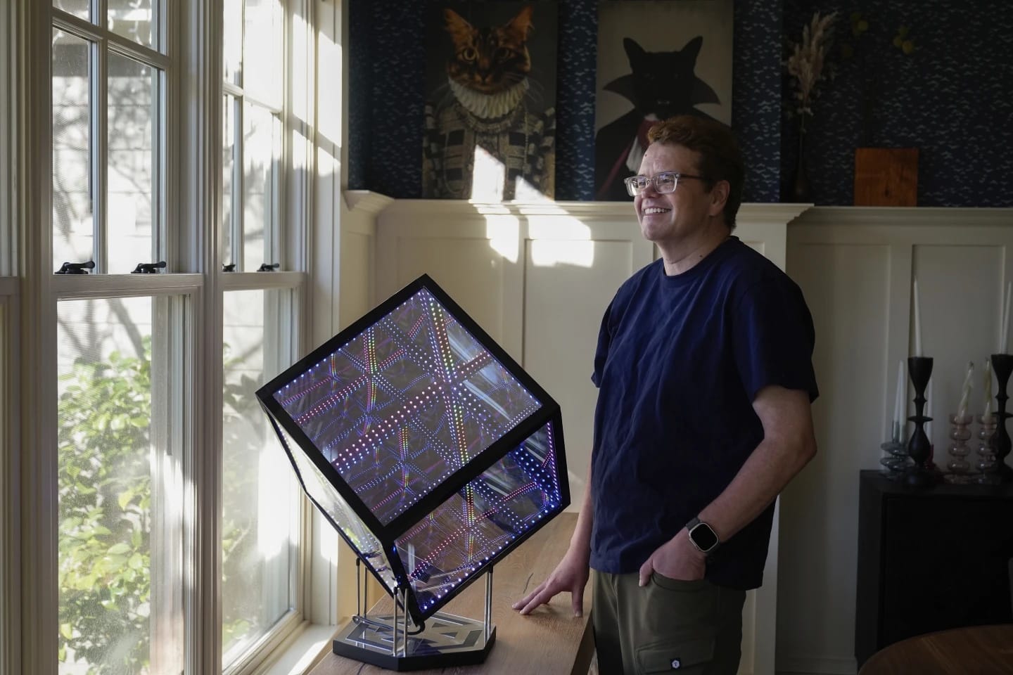 Jeff Atwood inside home standing and looking out a window with a Hypercube (these are awesome) and renaissance era portraits of their two cats, Scout and Blake