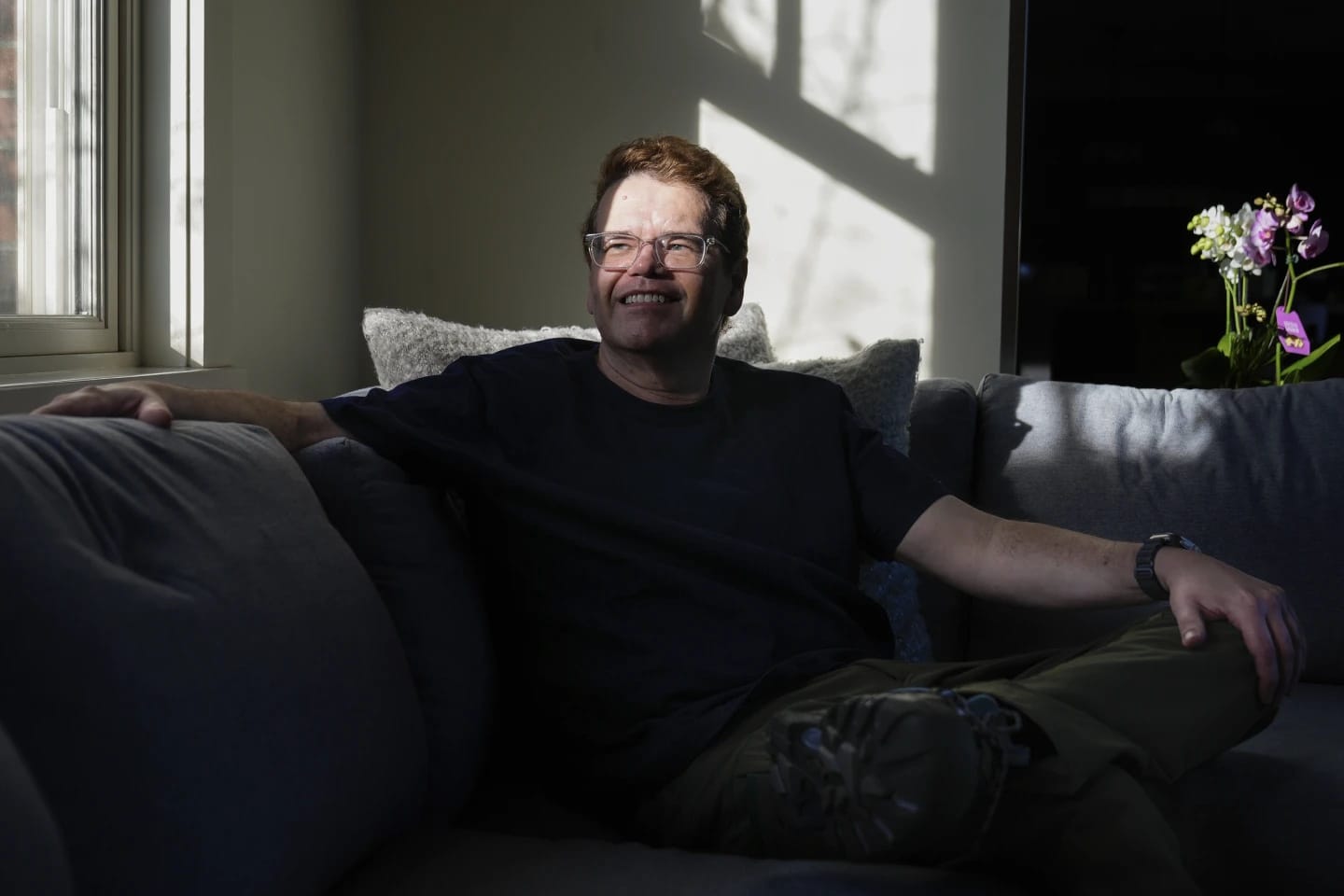 Jeff Atwood inside home sitting on a couch, legs crossed, both arms resting outward, half in light, half in shadow