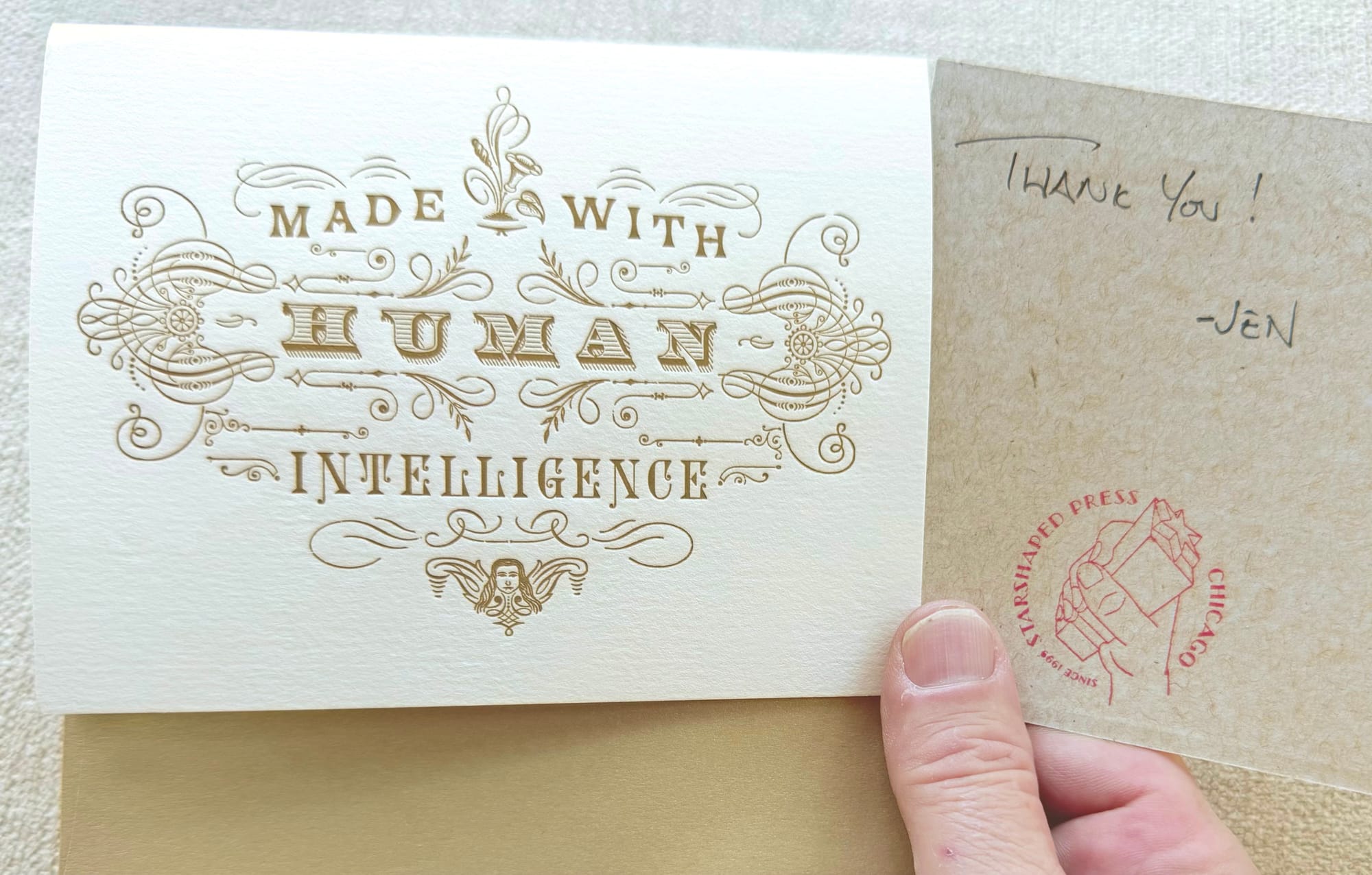 small folded postcard from Starshaped Press in Chicago, IL with gold envelope and gold ink -- "Made with human intelligence" in complex letterpress layout, alongside a small card with "Thank you! - Jen" handwritten on it, and the starshaped press logo stamped on it.
