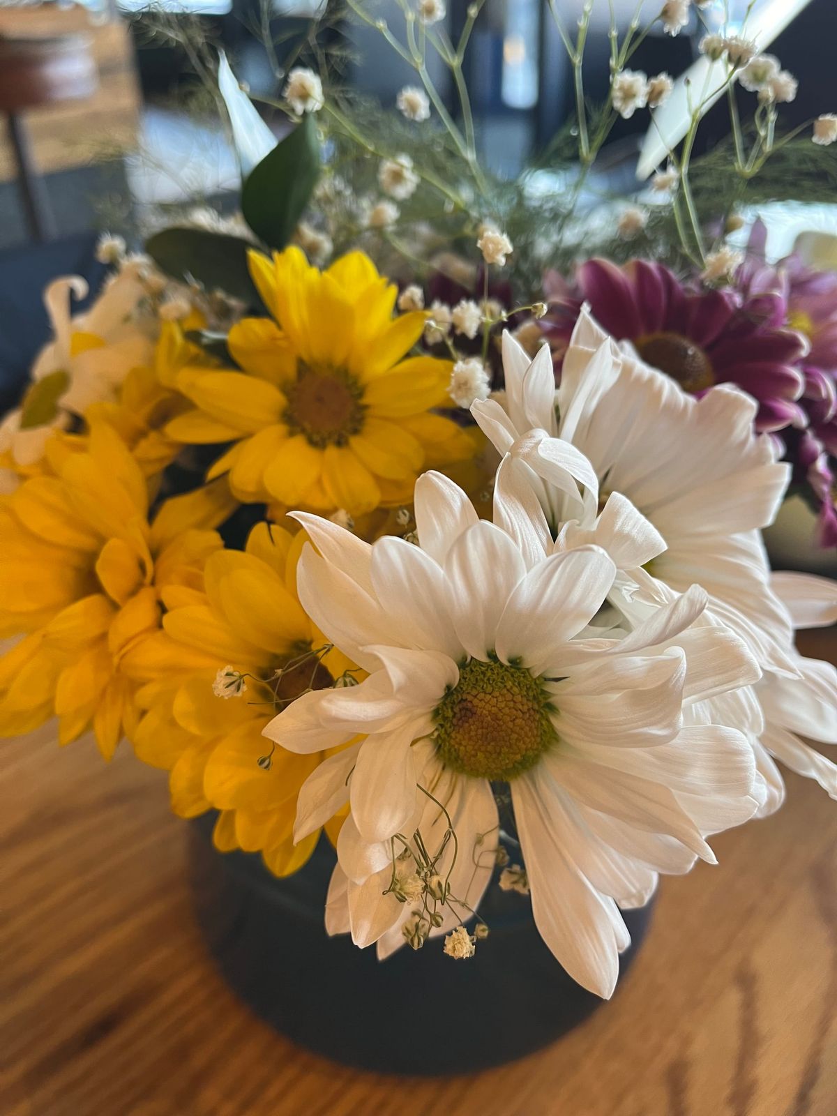 A bouquet of yellow, white, and purple daisies and baby's breath in a Le Creuset vase on a butcher block table.