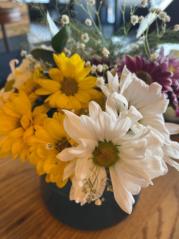 A bouquet of yellow, white, and purple daisies and baby's breath in a Le Creuset vase on a butcher block table.