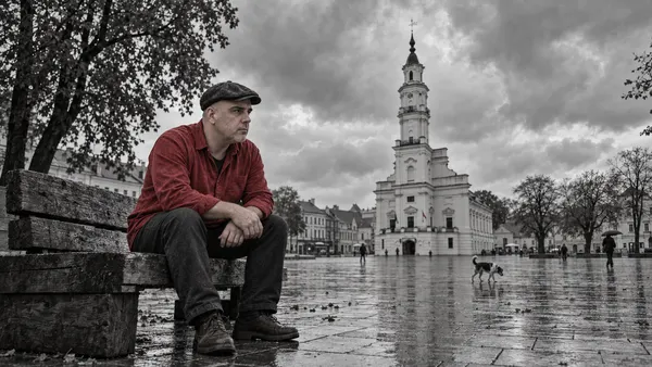 Evaldas su kaimiška kepure ir raudonais marškiniais sėdi ant grubaus medinio suoliuko lietingoje Kauno Rotušės aikštėje; virš rotušės slenka debesys, netoliese eina šuo.