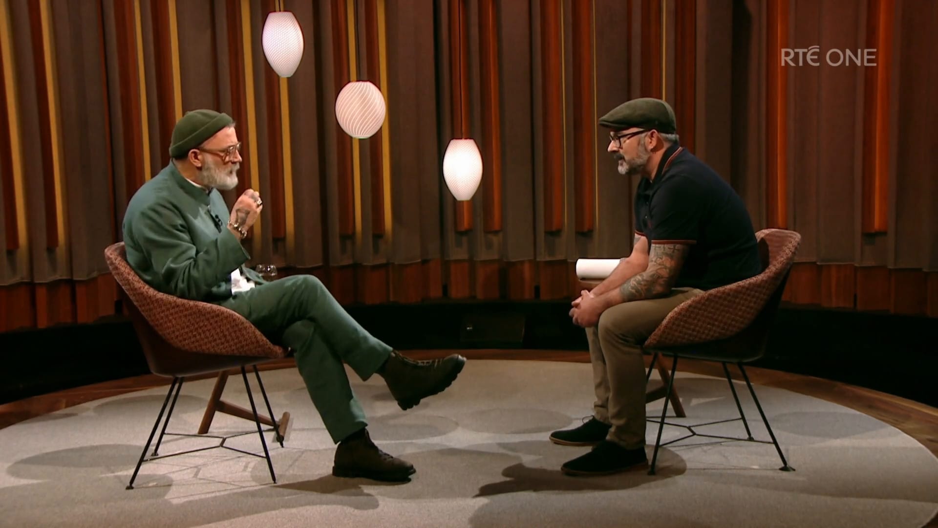Two men sit on chairs facing each other in a TV studio for an interview. The interviewer is intrigued by the guest. 