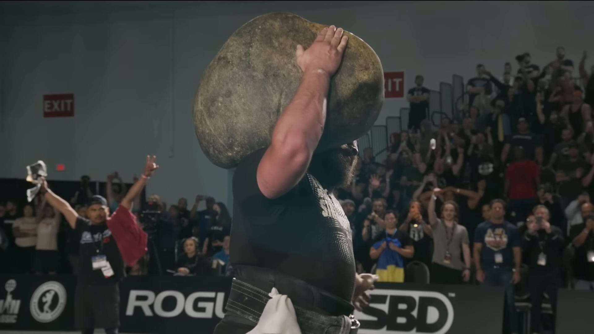 Martins Licis has a huge stone balanced on his right shoulder with his left hand held out to the side to show his control.