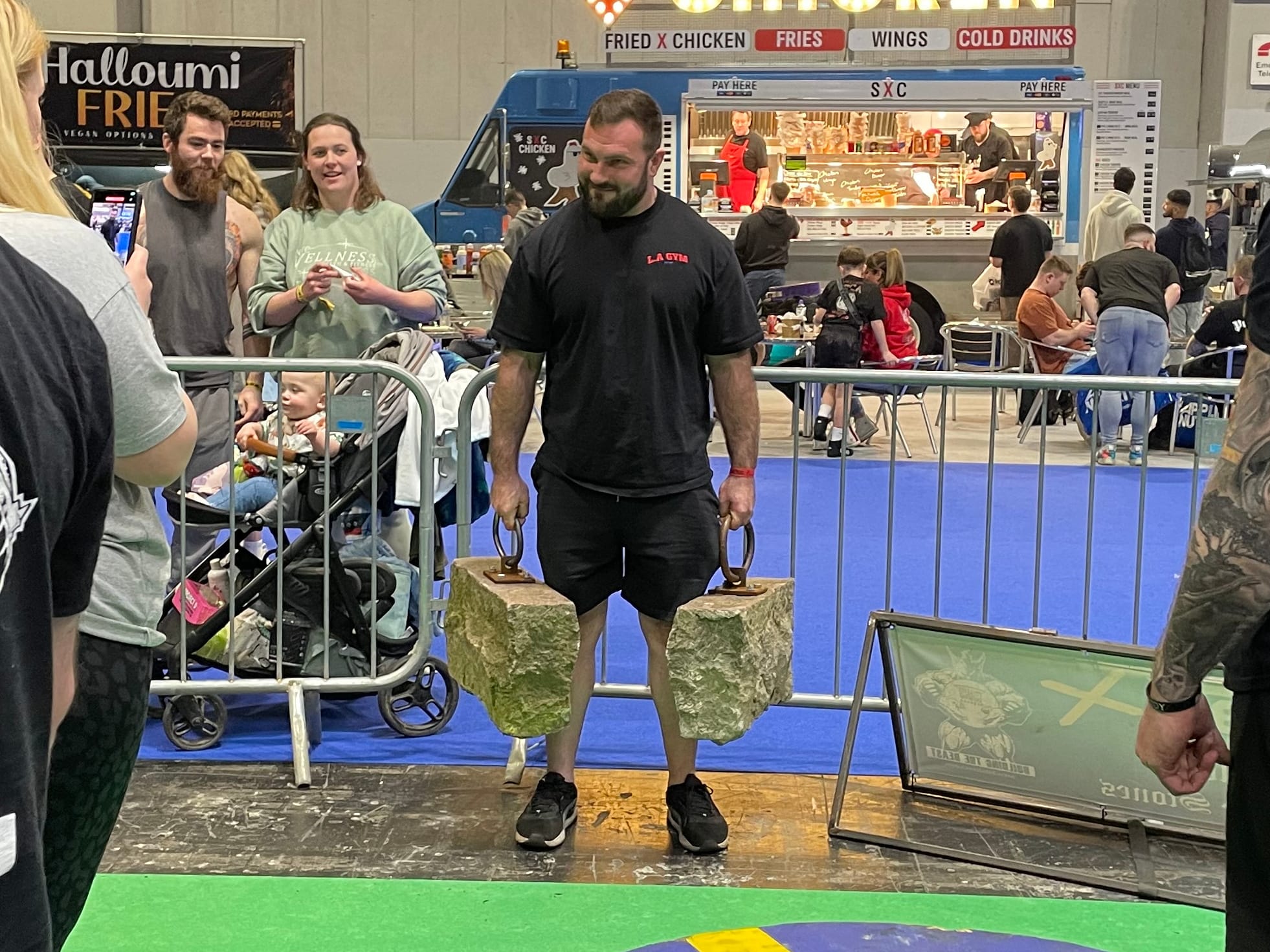 A person is stood inside a gated area. He's holding two metal rings in his hands attached to rough stones that scrape his shins.