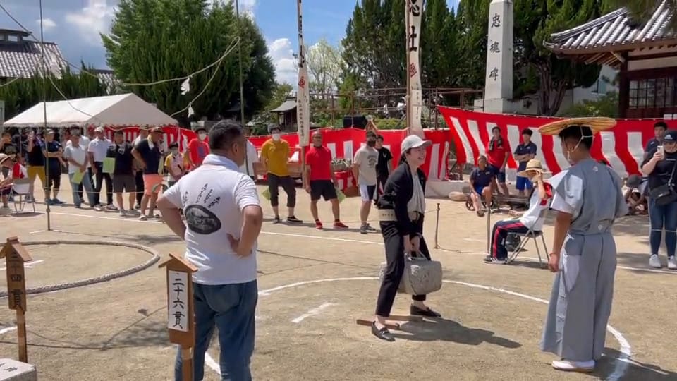 A Japanese politician wearing a suit and a weightlifting belt lifts a stone using its harness.