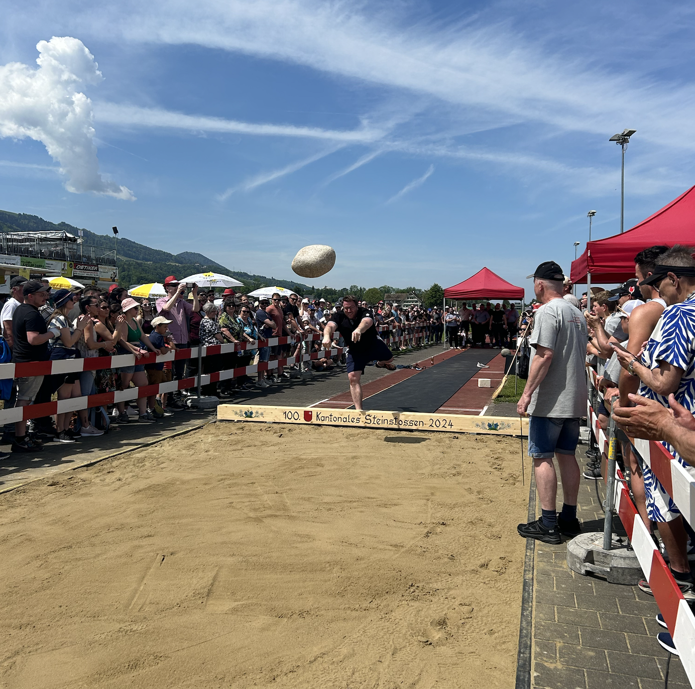 A man is fully extended at a toe beam with his arms outstreched. A large stone is flying through the air towards a sandpit.