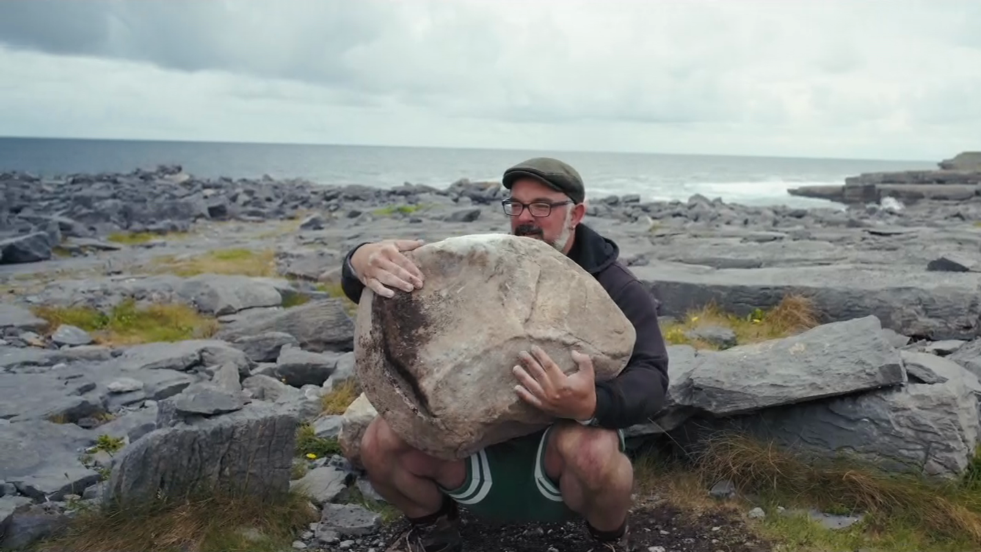 David Keohan is squatting down with the pinkish Inish More Stone resting on his lap, he's preparing to stand up with the stone.