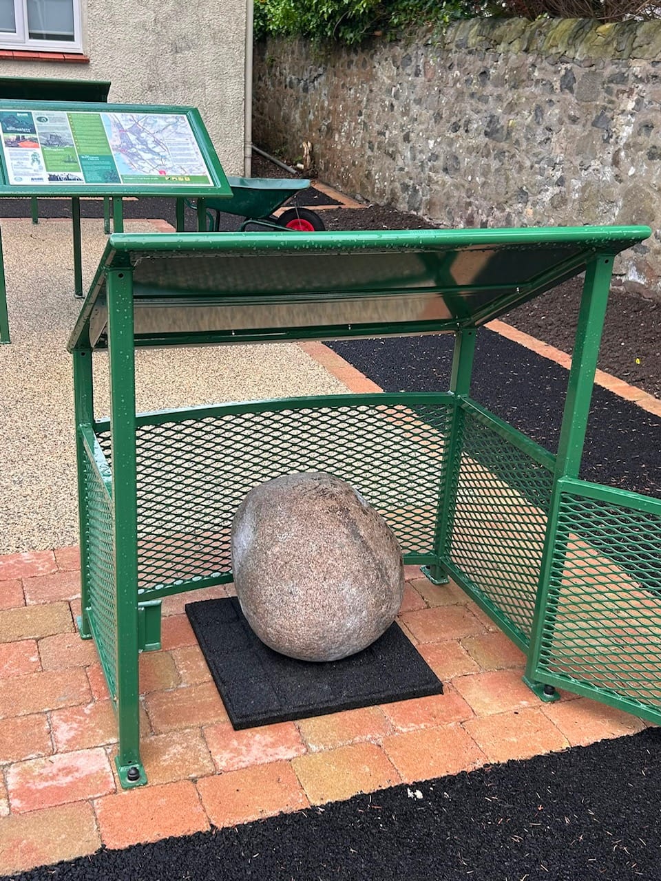 The Old Loans Stone sitting on a black rubber tile mat inside an open green cage used to store it at the Loans Village Hall garden..