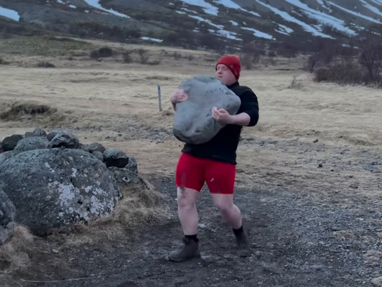 A man wearing a red beanie carries a huge basalt slab on his chest.
