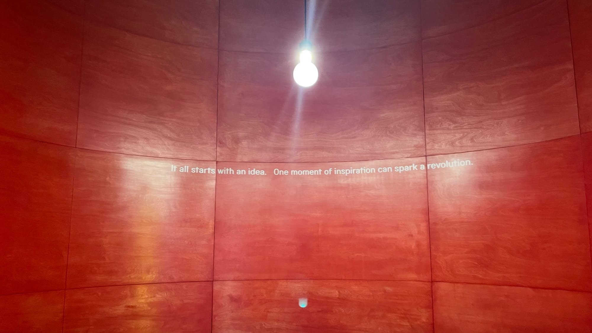 A quote is written on a red wall with a bright lightbulb hanging from the ceiling. "It all starts with an idea.  One moment of inspiration can spark a revolution".