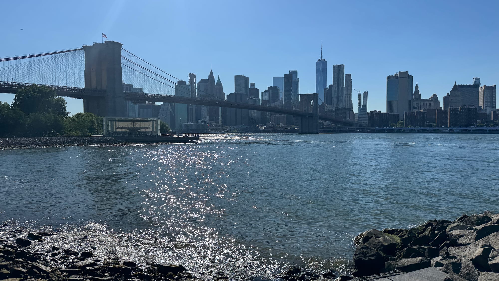 A view of Brooklyn Bridge, Jane's Carousel, and Manhattan from the stony shore of Brooklyn Bridge Park.
