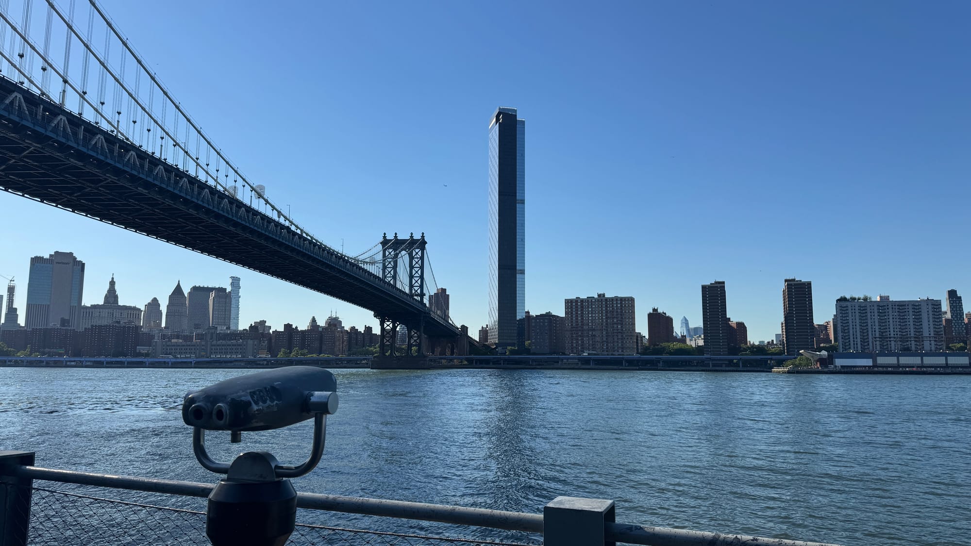 A pair of binoculars in Brooklyn Bridge Park pointing across the East River, facing One Manhattan Square. Manhattan Bridge is also visible on the side.