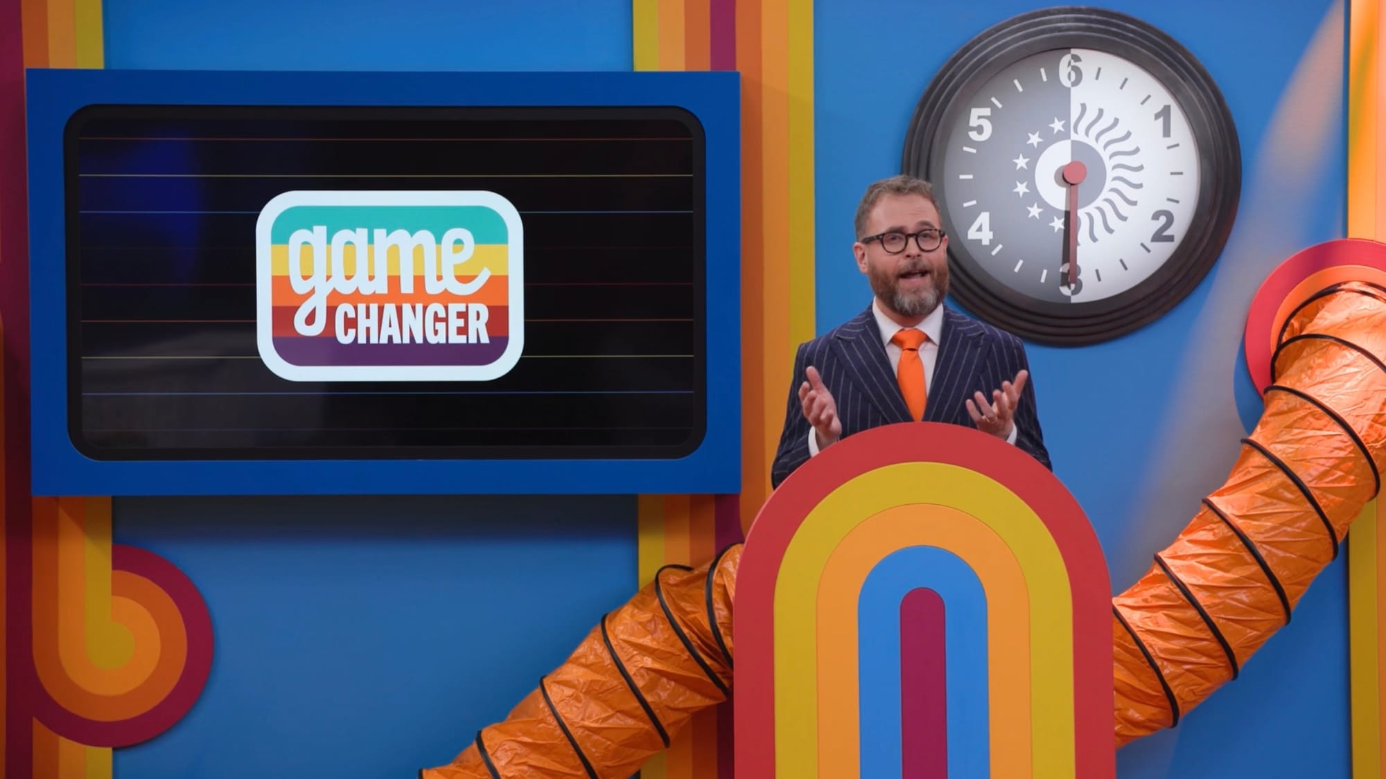 A screenshot from Game Changer S8Ep3: Night Shift. The host, Sam Reich, is standing on his podium, with a clock behind him going up to 6, and a dark mode Game Changer logo.