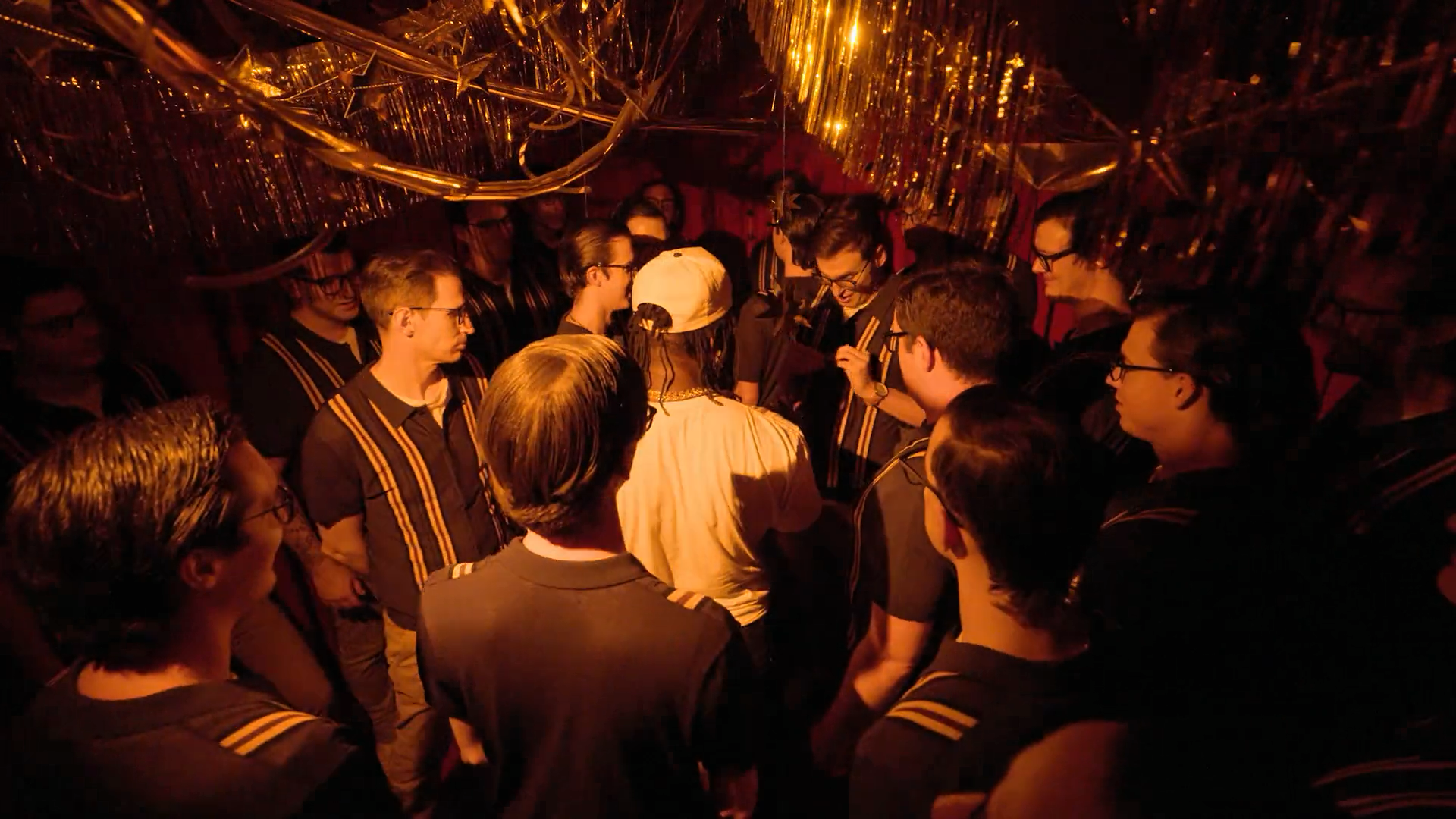 A screenshot from Game Changer S8Ep05, "Count the Rice". Ify Nwadiwe is in a harshly lit red room with Grant O'Brien, and over 20 people who look similar to Grant O'Brien.