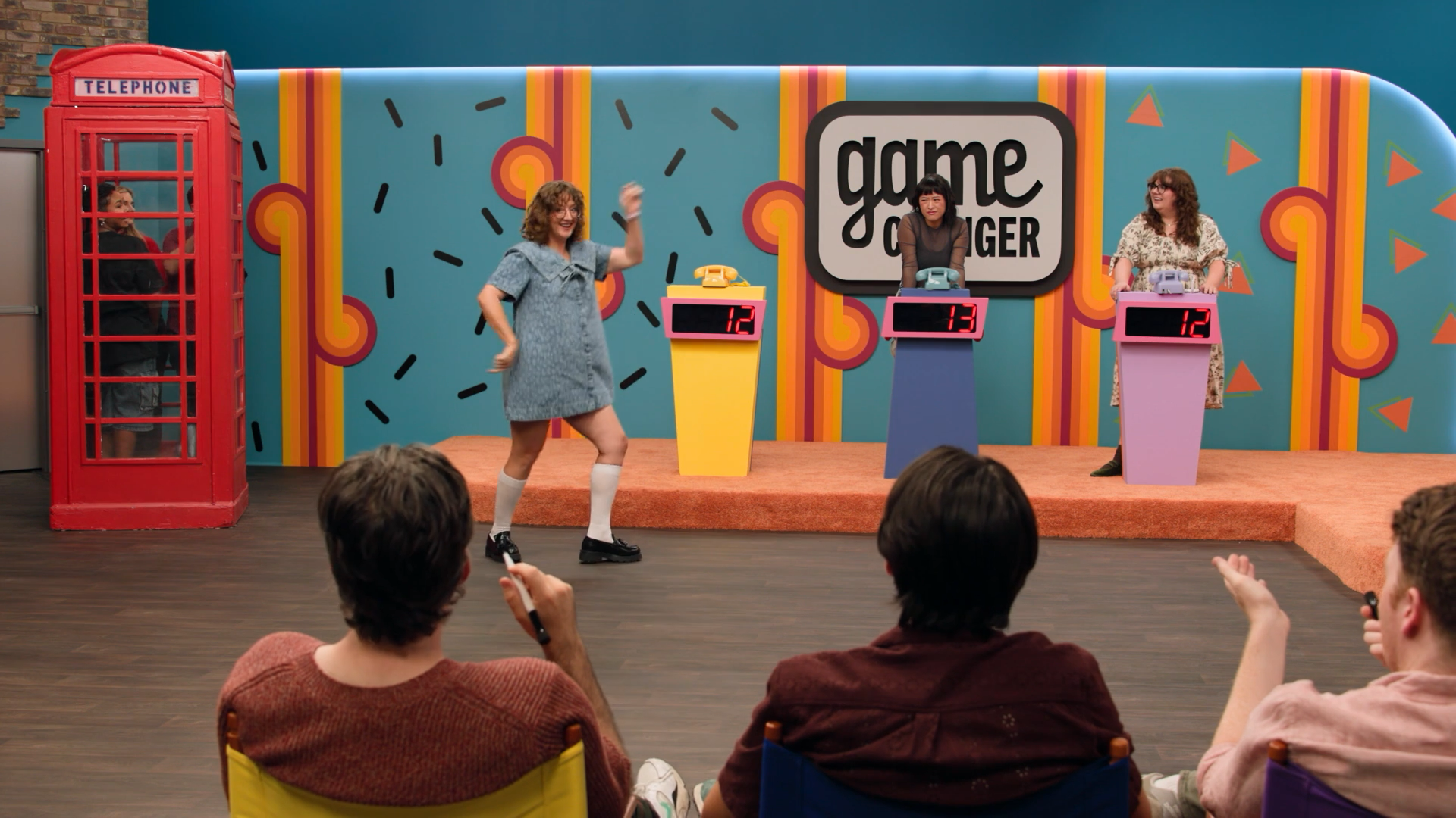 A screenshot from Game Changer S8Ep06, "Noise Some Makes", featuring Jess Ross performing a prompt, Lily Du and Jordan Myrick on the other podiums, the Noise Boys watching them, and the artists crammed in a phone booth.