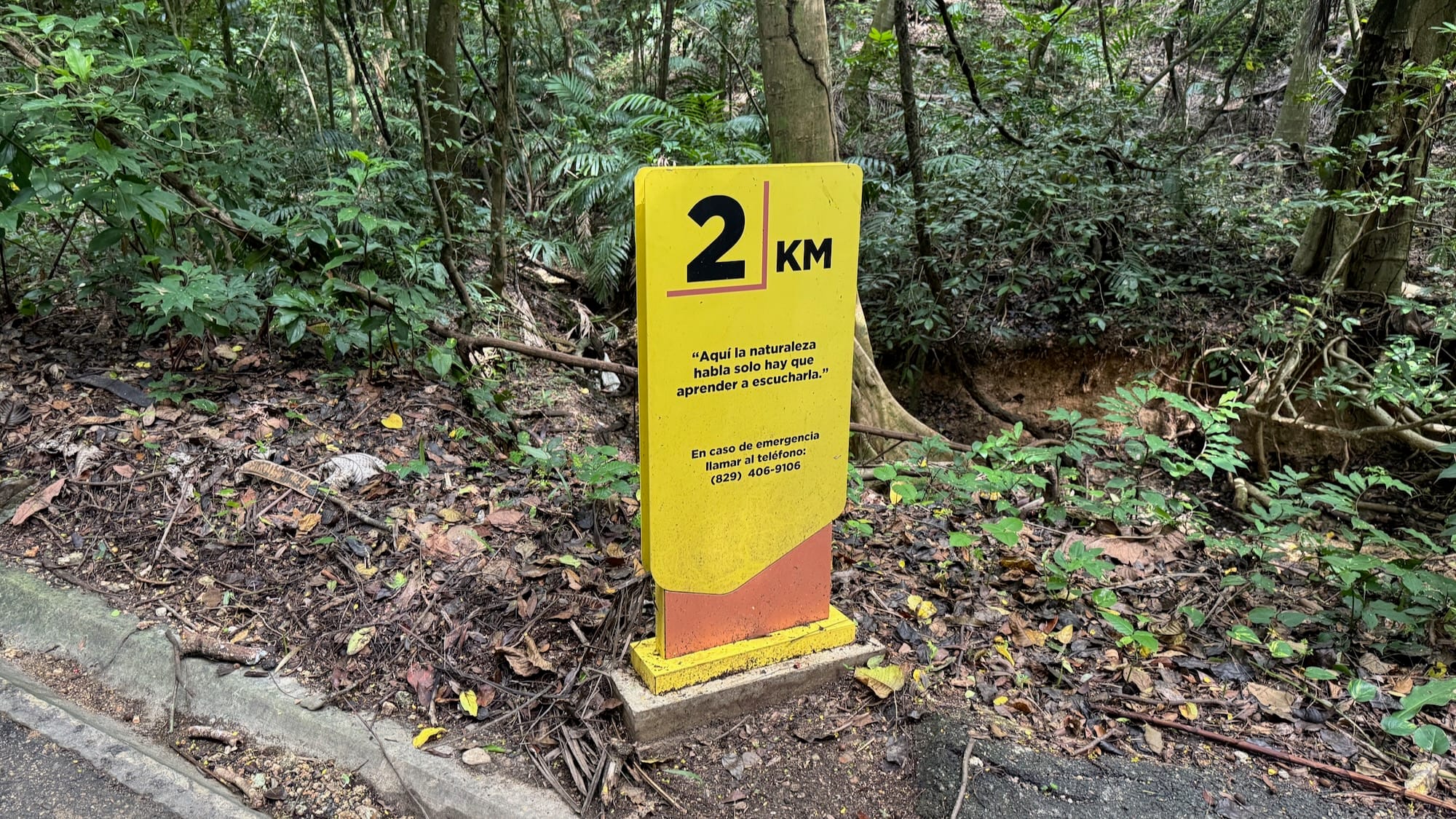 Un letrero marcando 2 kilometros en una caminata del Jardín Botánico Nacional de Santo Domingo. Dice "Aquí la naturaleza habla solo hay que aprender a escucharla." / A sign marking 2 kilometres in the Santo Domingo Botanical Garden.