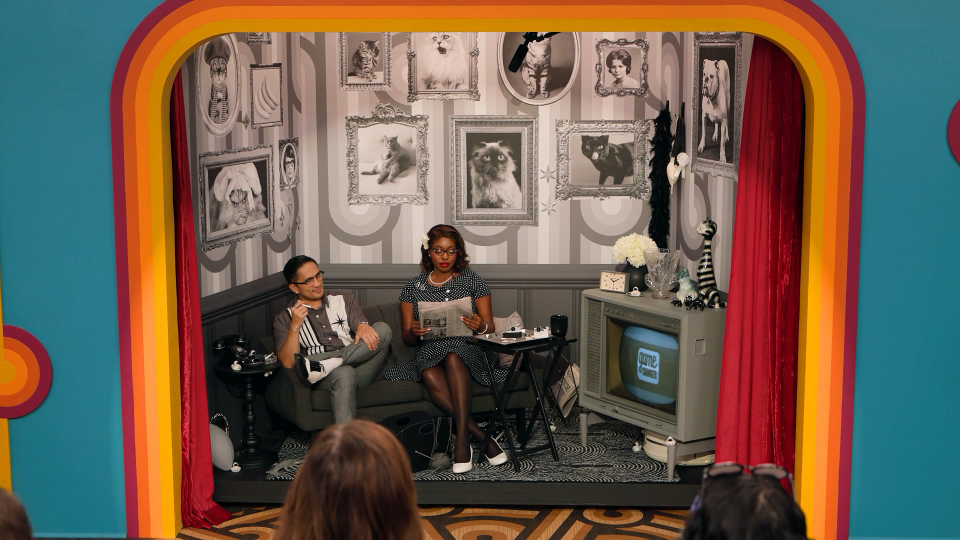 A screenshot of Game Changer S8Ep9, "I Spy", showing their 1950s styled tableau. A couple is sitting in a black and white room wearing black and white clothes, one is smoking a cigarette while the other reads a newspaper. There's cake on the table.