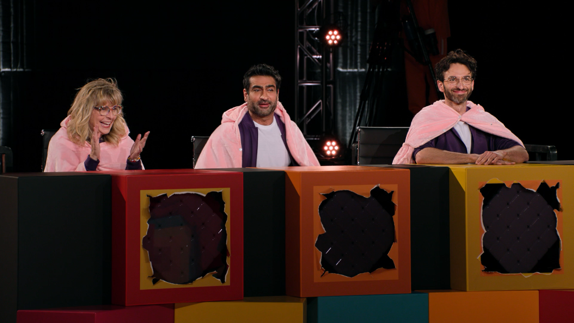 A screenshot of Game Changer S8Ep10, "Last Talent Standing". The three judges, Maria Bamford, Kumail Nanjiani, and Gianmarco Soresi, are sitting in their podiums with purple jumpsuits and pig coats. Their podiums are bright-coloured paper boxes with holes punched out to show their screens.