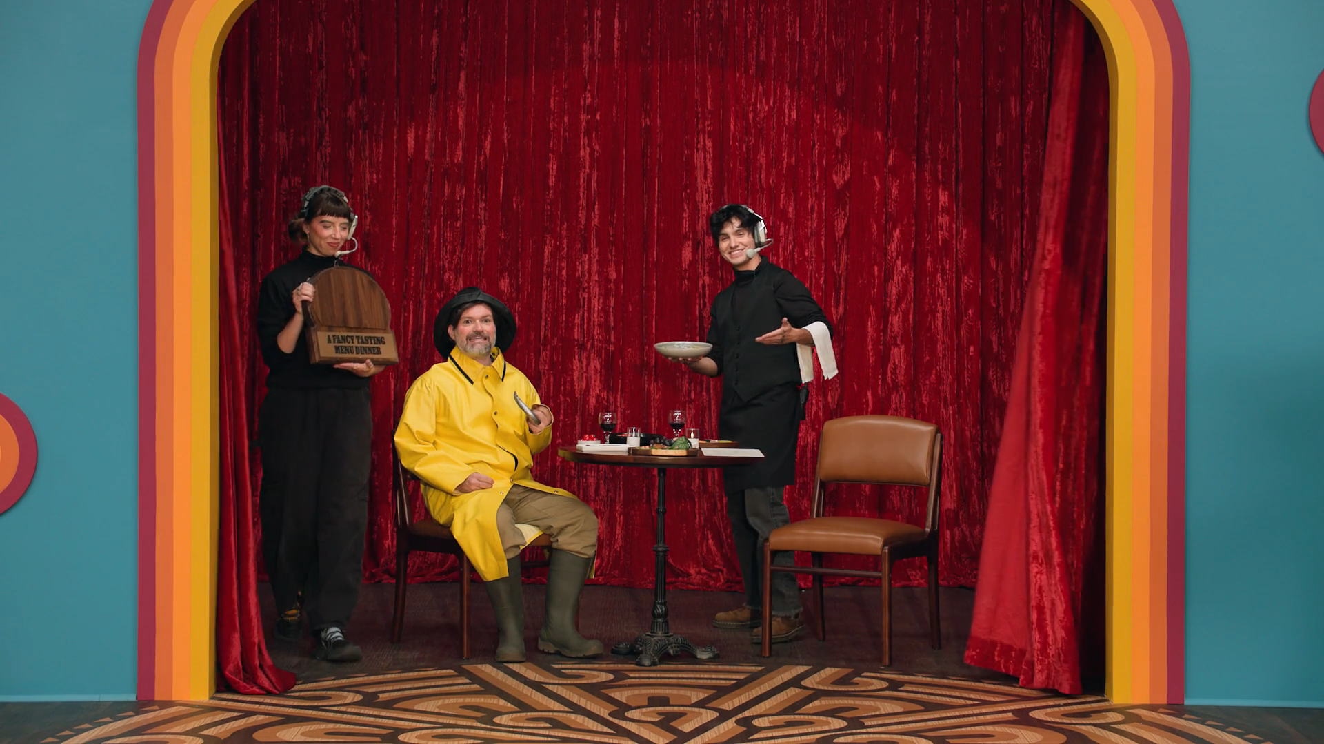 A screenshot from Game Changer S8Ep08, "What's The Catch". Ash Kron and Nico Romero presenting the prize of A Fancy Tasting Menu Dinner. Josh Ruben is also there, posing in a chair as a creepy fisherman.