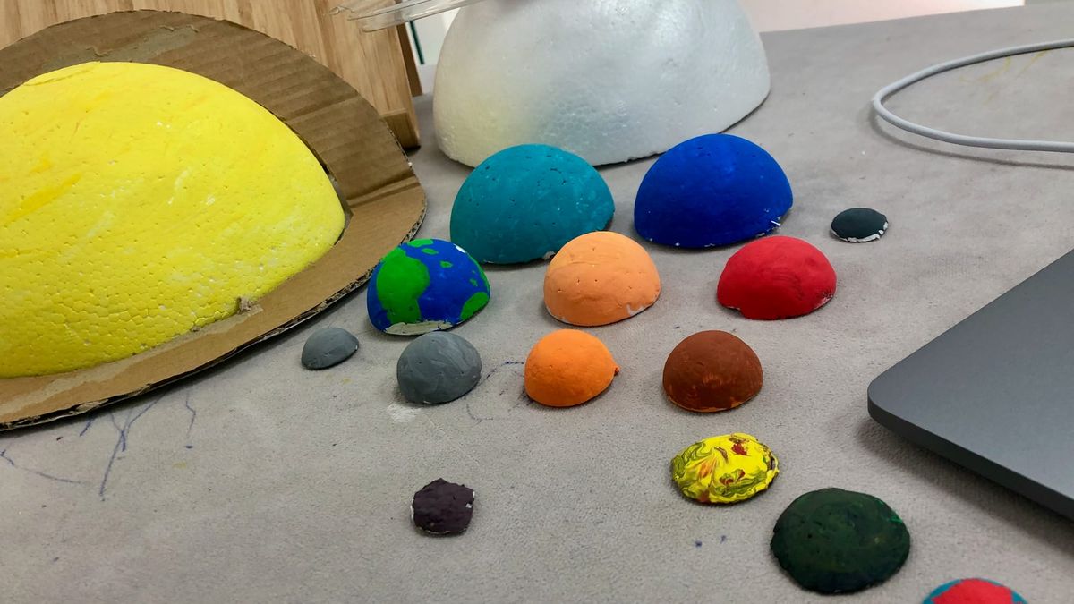 An image showing a bunch of foam semispheres coloured to resemble planets and major moons of the Solar System on a table. A brown loop of cardboard is Saturn's rings. Jupiter isn't painted.