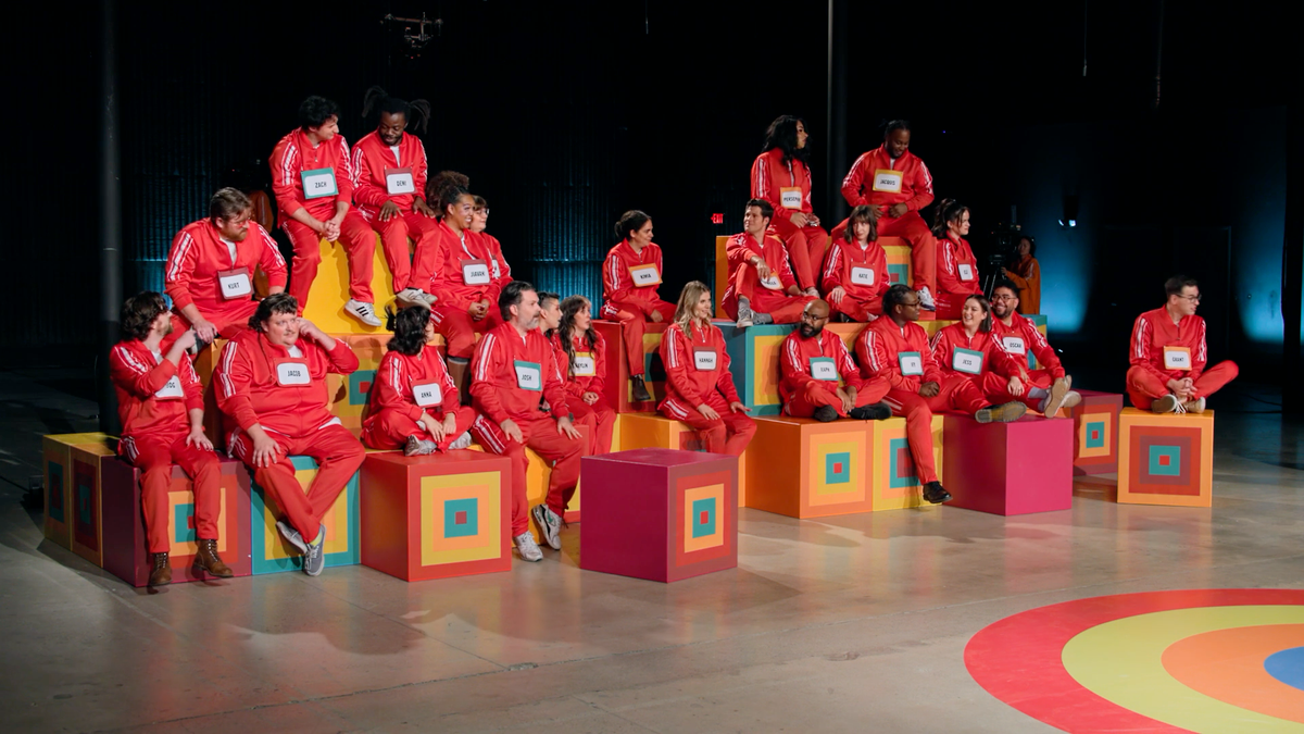 A screenshot of Game Changer S8Ep10, "Last Talent Standing". 23 contestants are sitting in a bunch of boxes, all wearing bright red jumpsuits and name tags.