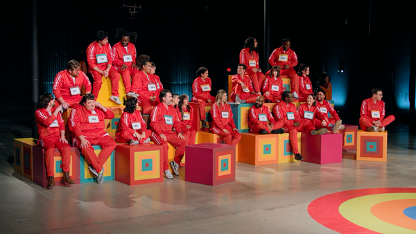 A screenshot of Game Changer S8Ep10, "Last Talent Standing". 23 contestants are sitting in a bunch of boxes, all wearing bright red jumpsuits and name tags.