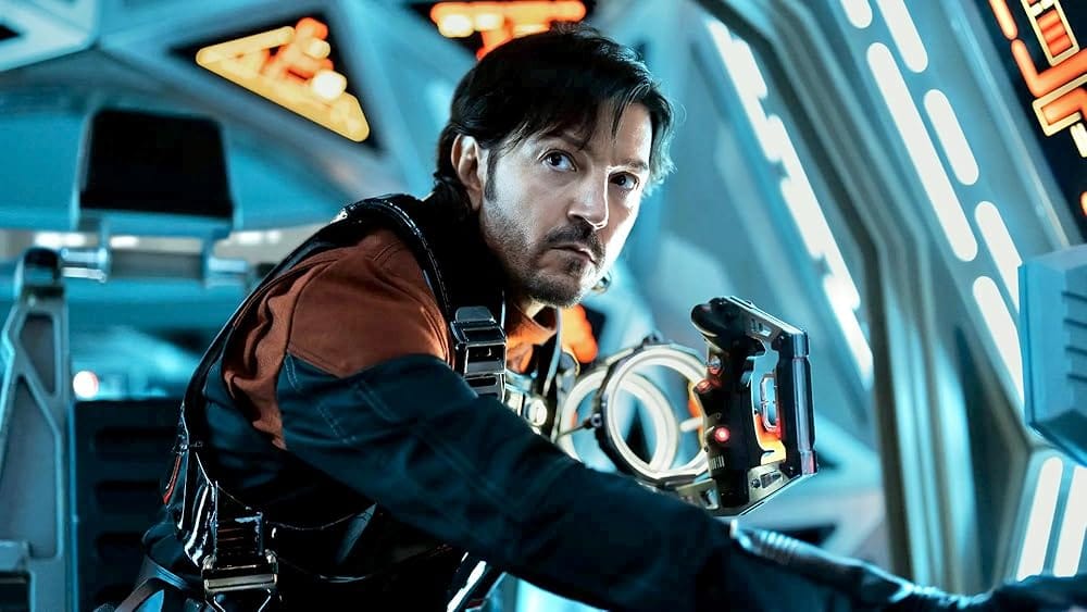 A man in a orange and black uniform sits in a mostly white spaceship.