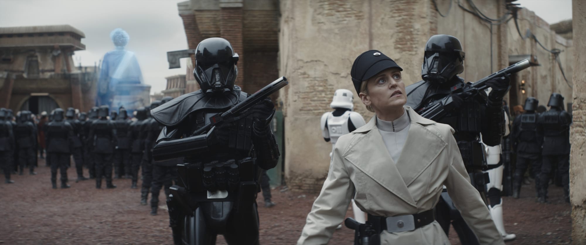 A woman in a white uniform is flanked by two troopers in black armor as they look for someone. Behind them a holoprojection is about to start some shit.