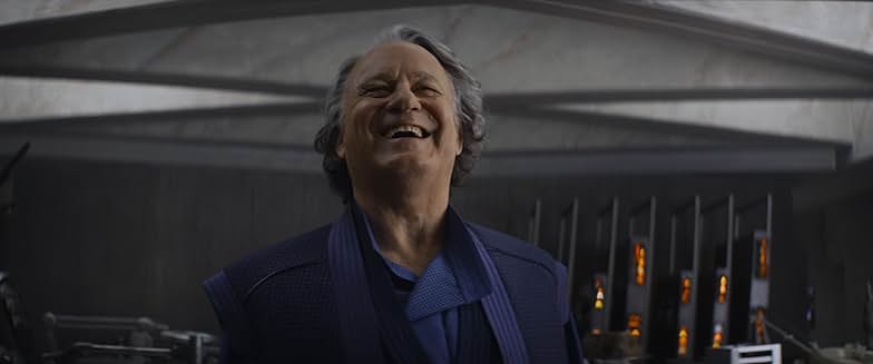 An older man in a blue suit stands in a workshop and laughs a hearty laugh.