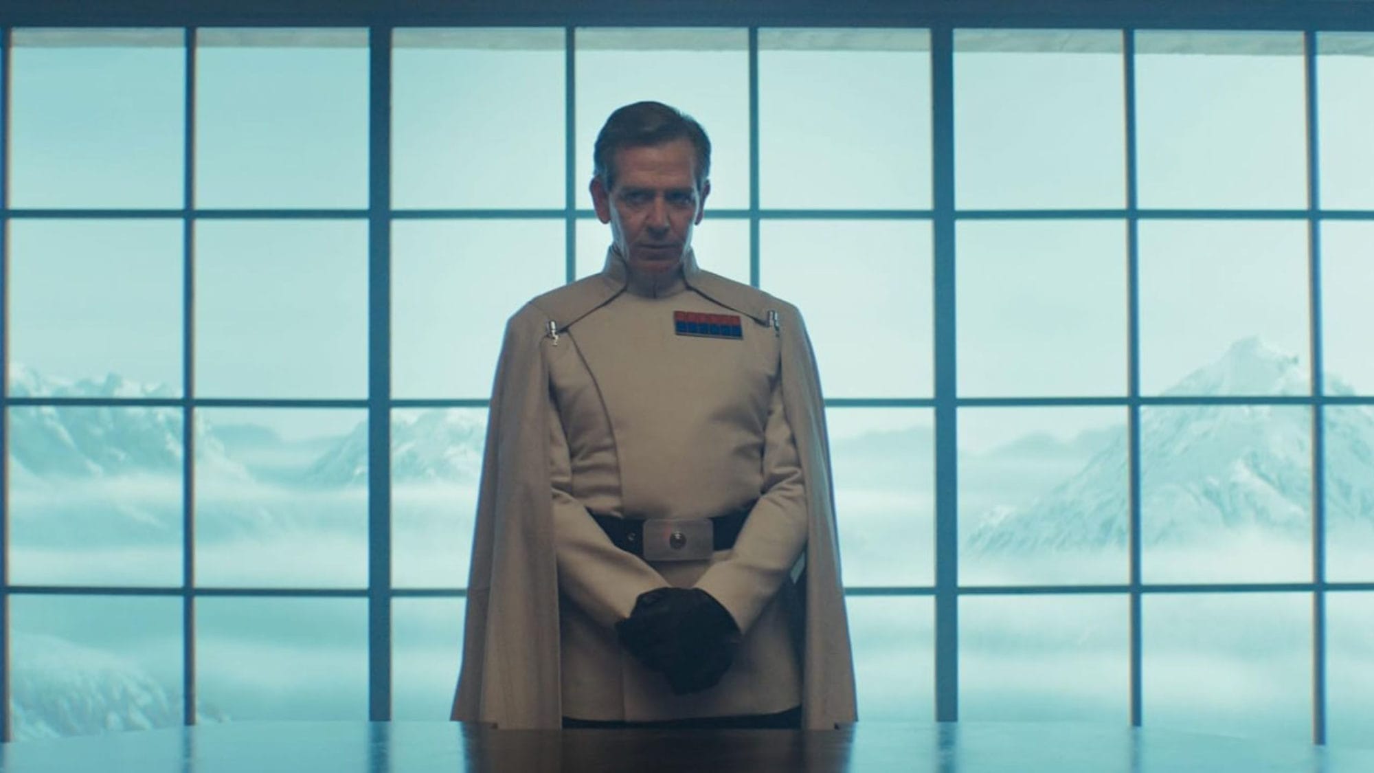 A man in a white uniform and a cape stands in front of a large window overlooking snow-capped mountains.