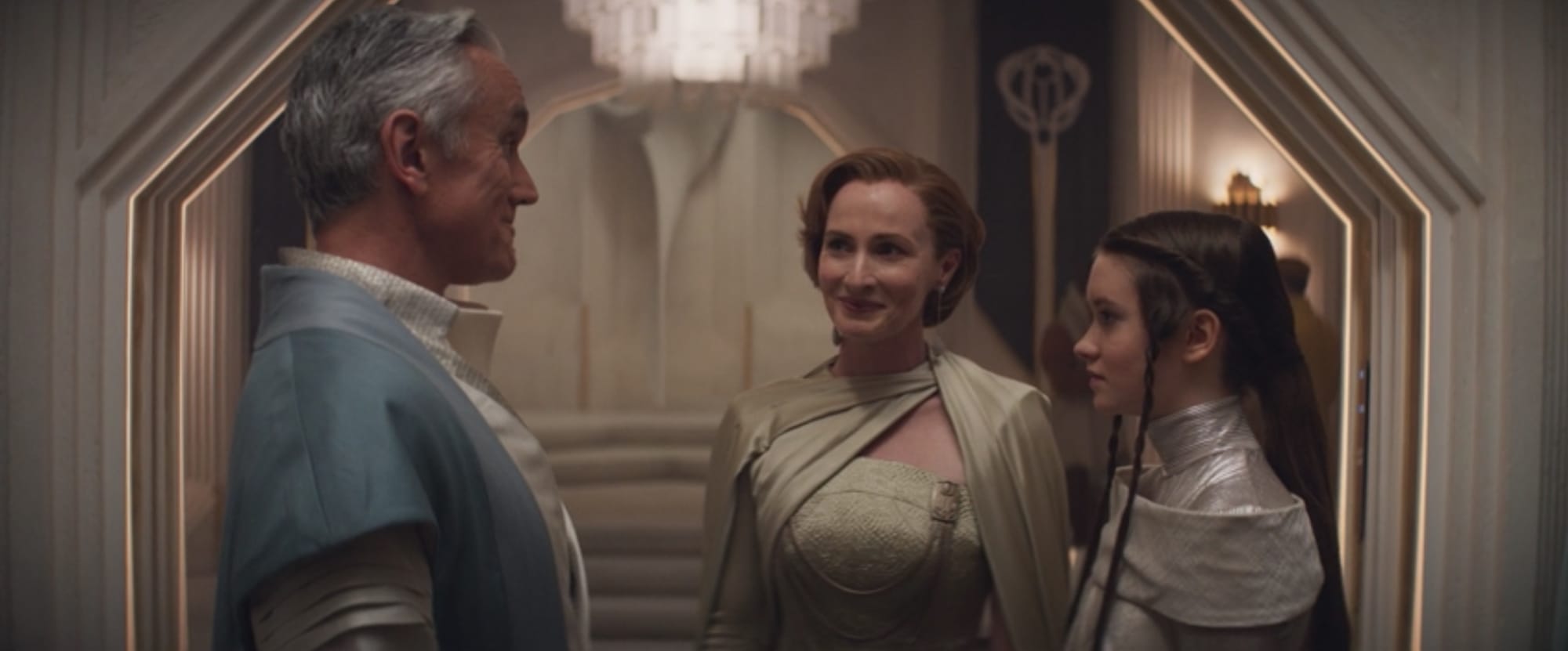 A taller man in a blue coat talks with a woman with red hair in a yellow dress and a teenage girl with brown hair in a silver a dress. They are in well appointed room in a large penthouse in Coruscant.