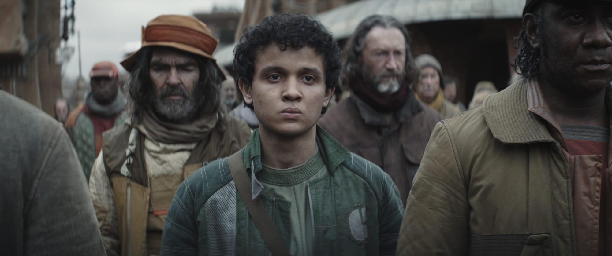A young man in a green coat stands amongst other men in brown and orange coats in a funeral procession.