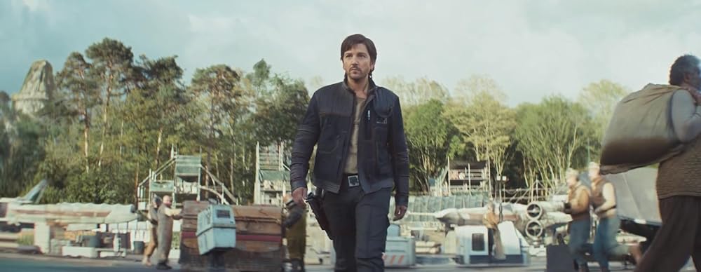 A man in a dark brown jacket and pants walks with purpose through a busy base on Yavin 4.