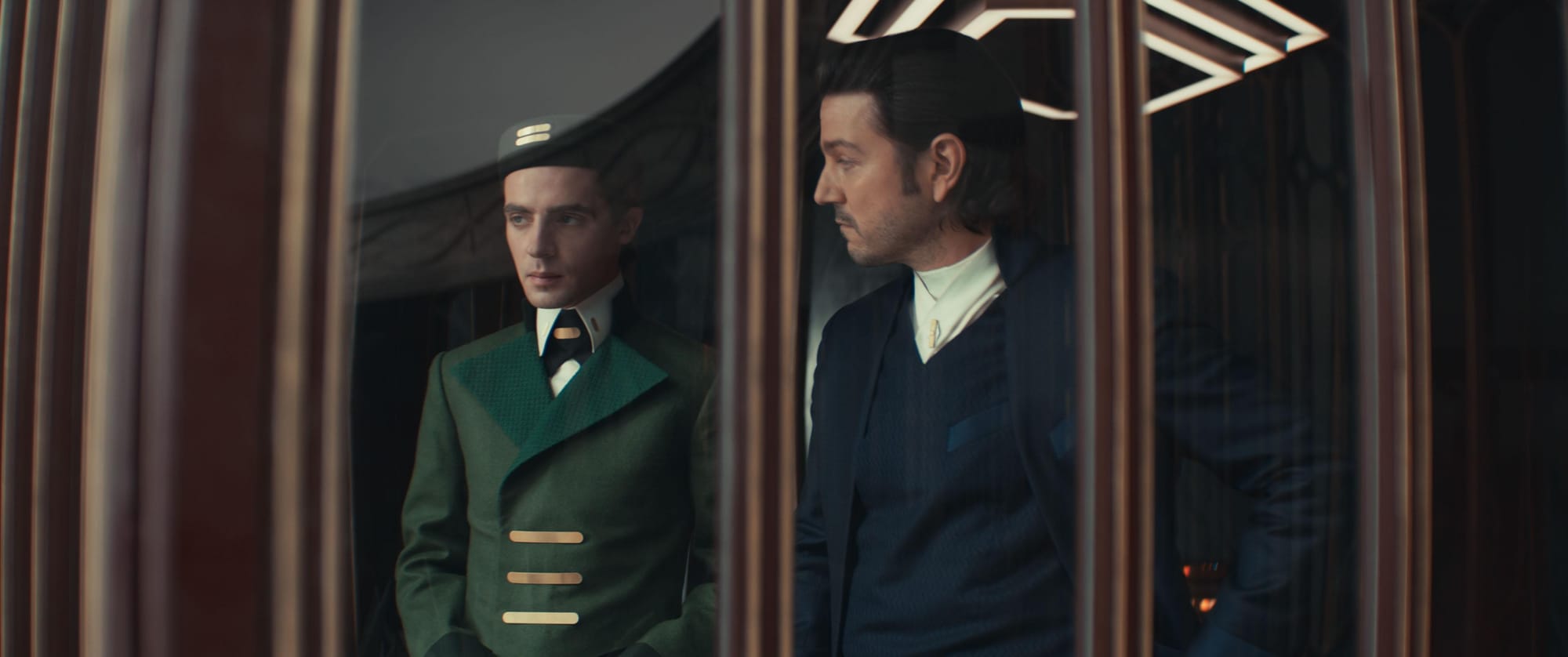  A man in a finely tailored blue suit talks to a bellhop in a green suit while both look out a hotel window